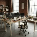 Estación de trabajo Hestia ambientada en oficina moderna estilo industrial con monitores y silla ergonómica.