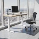 Escritorio de oficina regulable frente a ventanal con vista urbana junto a silla ergonómica negra y monitor.